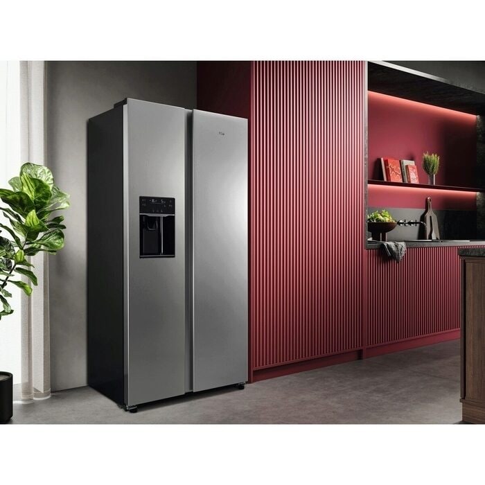 Moderne Küche mit einem freistehenden Side-by-Side-Kühlschrank in Edelstahloptik mit integriertem Wasserspender. Rechts daneben befindet sich eine rot gerippte Wandverkleidung mit offenen Regalen, dekoriert mit Büchern, Pflanzen und Küchenaccessoires. Links steht eine grüne Zimmerpflanze vor einer hellen Wand. Der Raum wirkt minimalistisch und stilvoll beleuchtet.