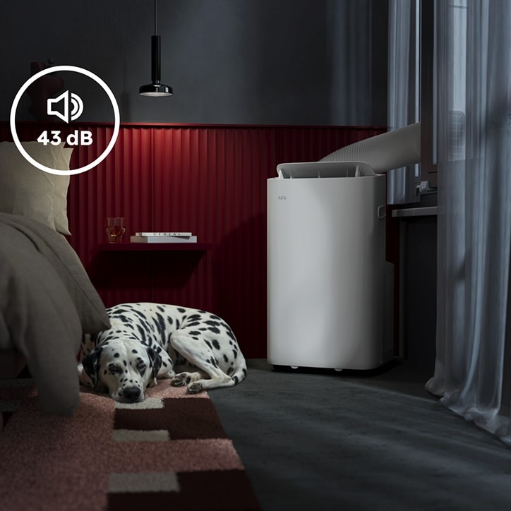 Leiser mobiler Klimagerät (43 dB) in einem modernen Schlafzimmer, neben einem Bett liegt ein schlafender Dalmatiner; das Gerät steht am Fenster mit Abluftschlauch.