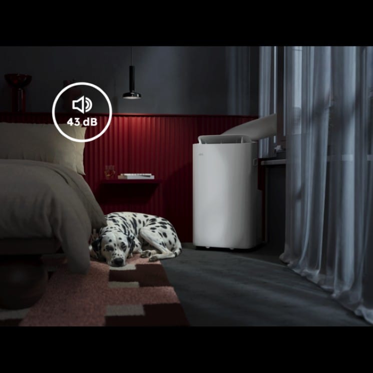 Leiser mobiler Klimagerät (43 dB) in einem modernen Schlafzimmer, neben einem Bett liegt ein schlafender Dalmatiner; das Gerät steht am Fenster mit Abluftschlauch.