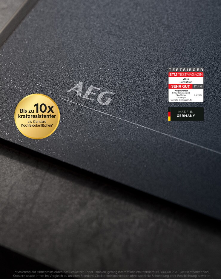 Das Bild zeigt eine dunkle, matte Kochfeldoberfläche mit dem AEG‑Logo in der Mitte. Links befindet sich ein goldener Rundaufkleber mit dem Hinweis ‚Bis zu 10x kratzresistenter als Standard‑Kochfeldoberflächen‘. Rechts sind zwei Label platziert: ein Testsiegel des ETM Testmagazins mit der Bewertung ‚SEHR GUT‘ sowie ein ‚Made in Germany‘‑Hinweis mit der deutschen Flagge. Die Oberfläche ist strukturiert und hochwertig.