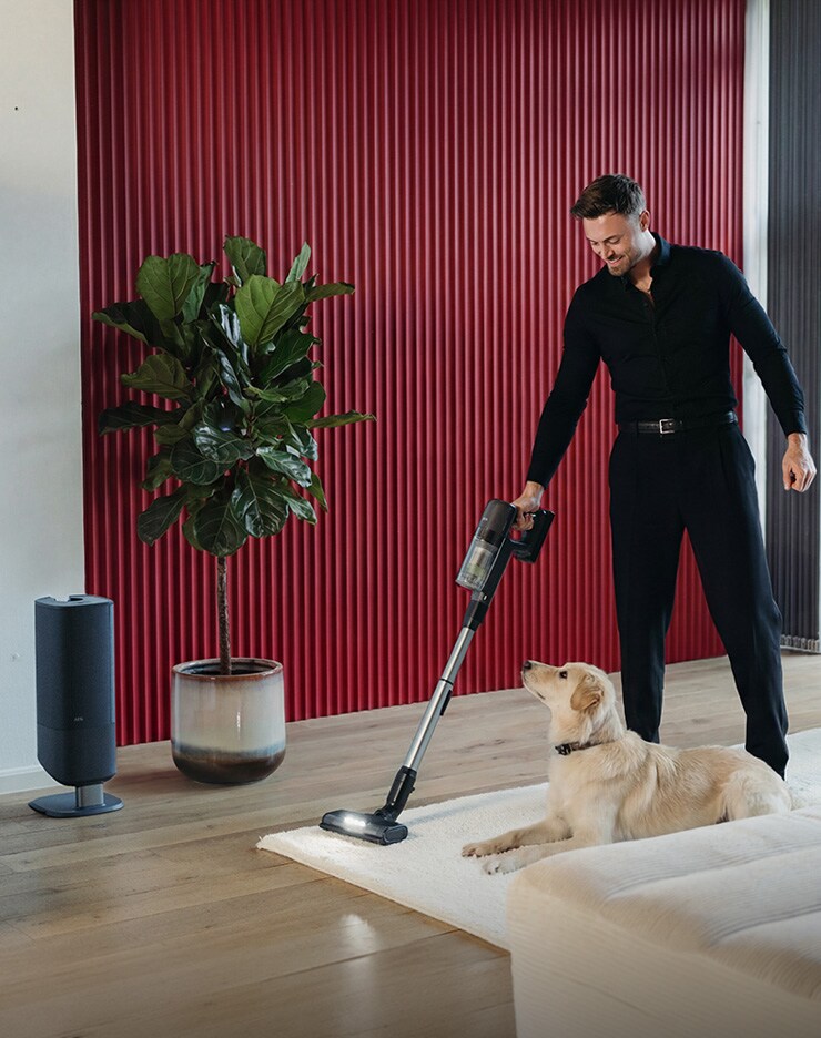 Person staubsaugt einen Teppich mit einem kabellosen Akku-Staubsauger, ein Hund liegt davor. Im Hintergrund steht die passende Staubsauger‑Station in einem modernen Wohnraum.