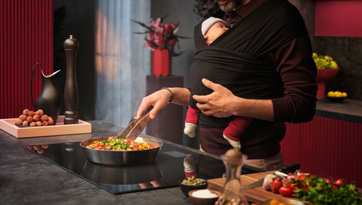 Ein Mann, der Essen auf einem Herd kocht, während er ein Baby in einer Babytrage auf der Brust trägt.