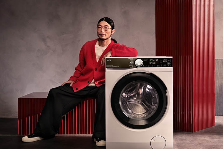 Mann in rotem Cardigan lehnt an AEG Waschmaschine der 9000er Serie vor grauem Hintergrund