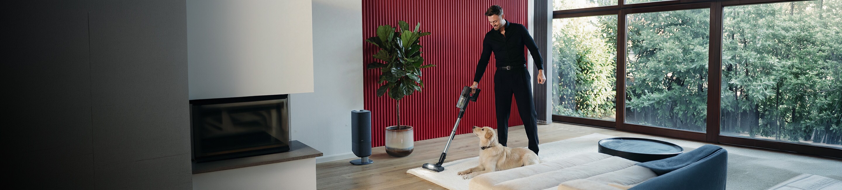 Person staubsaugt einen Teppich mit einem kabellosen Akku-Staubsauger, ein Hund liegt davor. Im Hintergrund steht die passende Staubsauger‑Station in einem modernen Wohnraum.